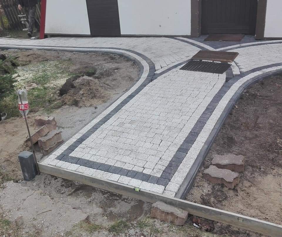 Nowo ułożona ścieżka z kostki brukowej w odcieniach szarości, z widocznym poziomem, butelką po napoju i stertą cegieł w pobliżu, prowadząca do drzwi budynku