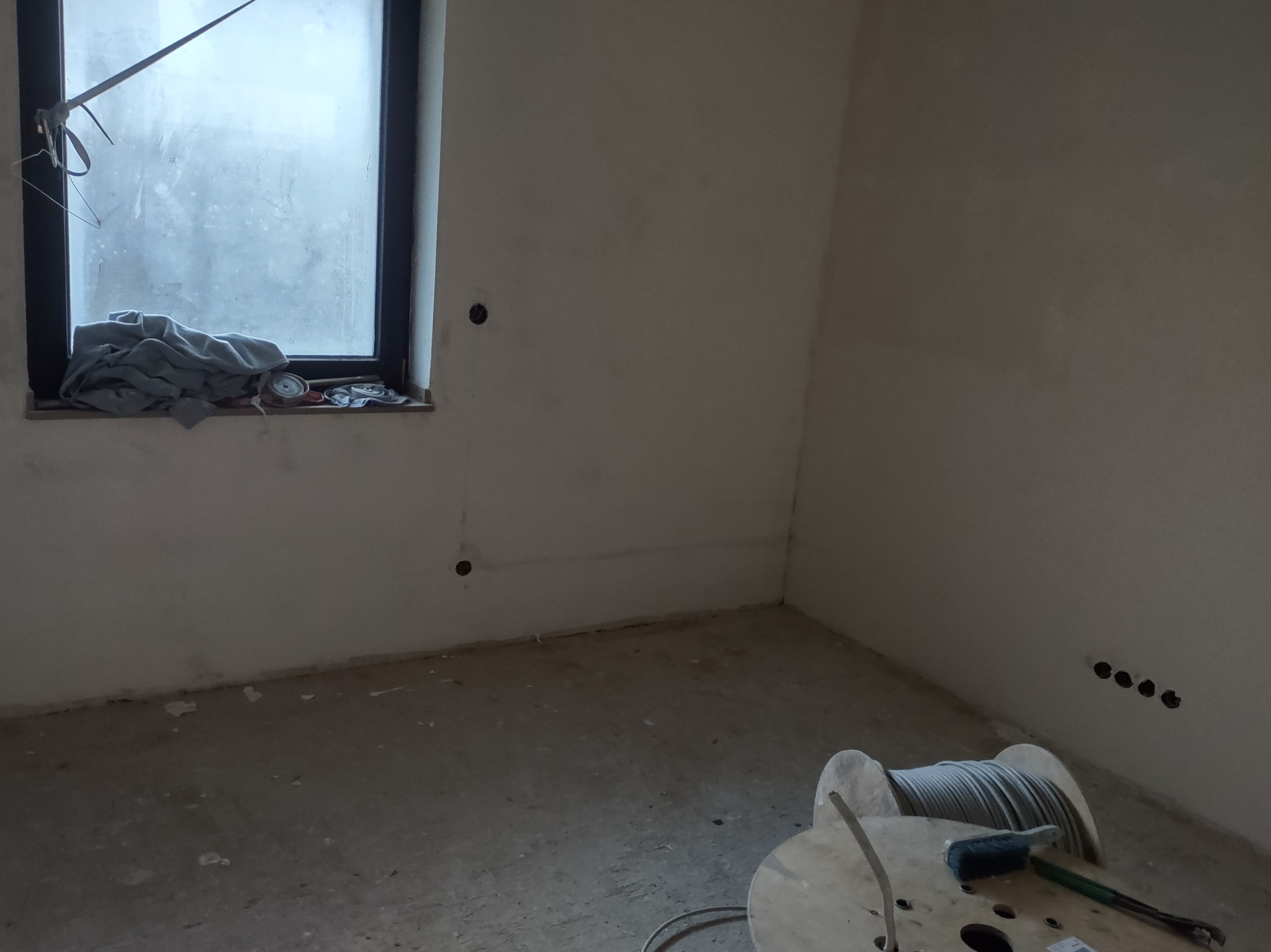 Surowe wnętrze pokoju w trakcie remontu: widoczne przewody na drewnianej szpuli, otwory w ścianie pod instalacje elektryczne, okno z ubraniami na parapecie.