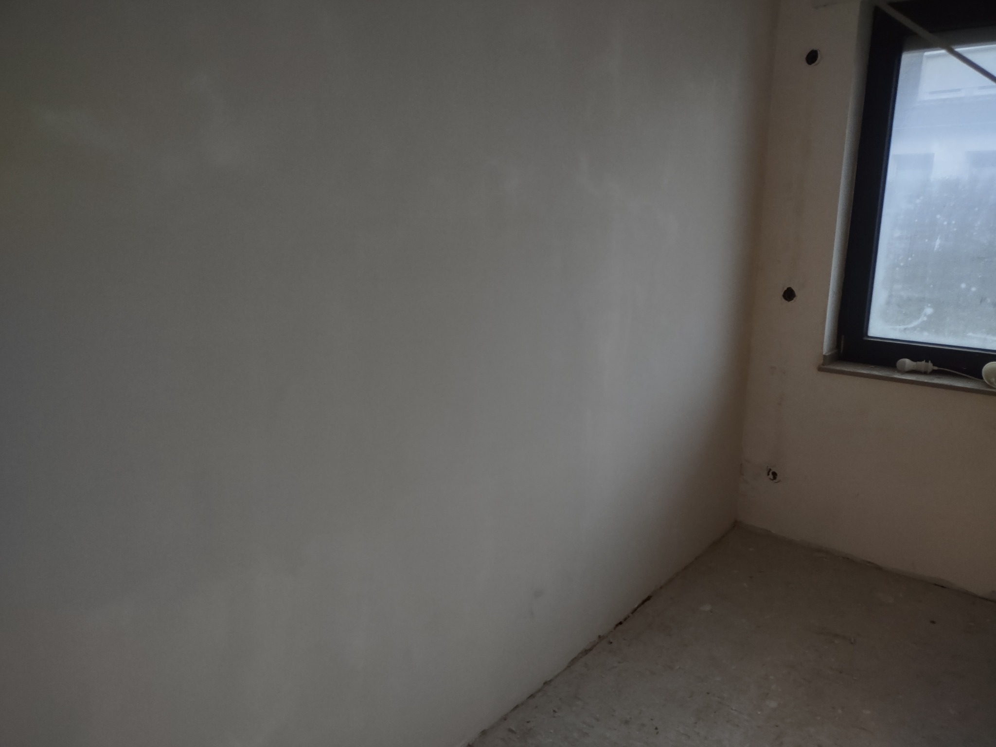 Surowa ściana po wyburzeniu, z widocznymi otworami po instalacjach elektrycznych i oknem w czarnej ramie.