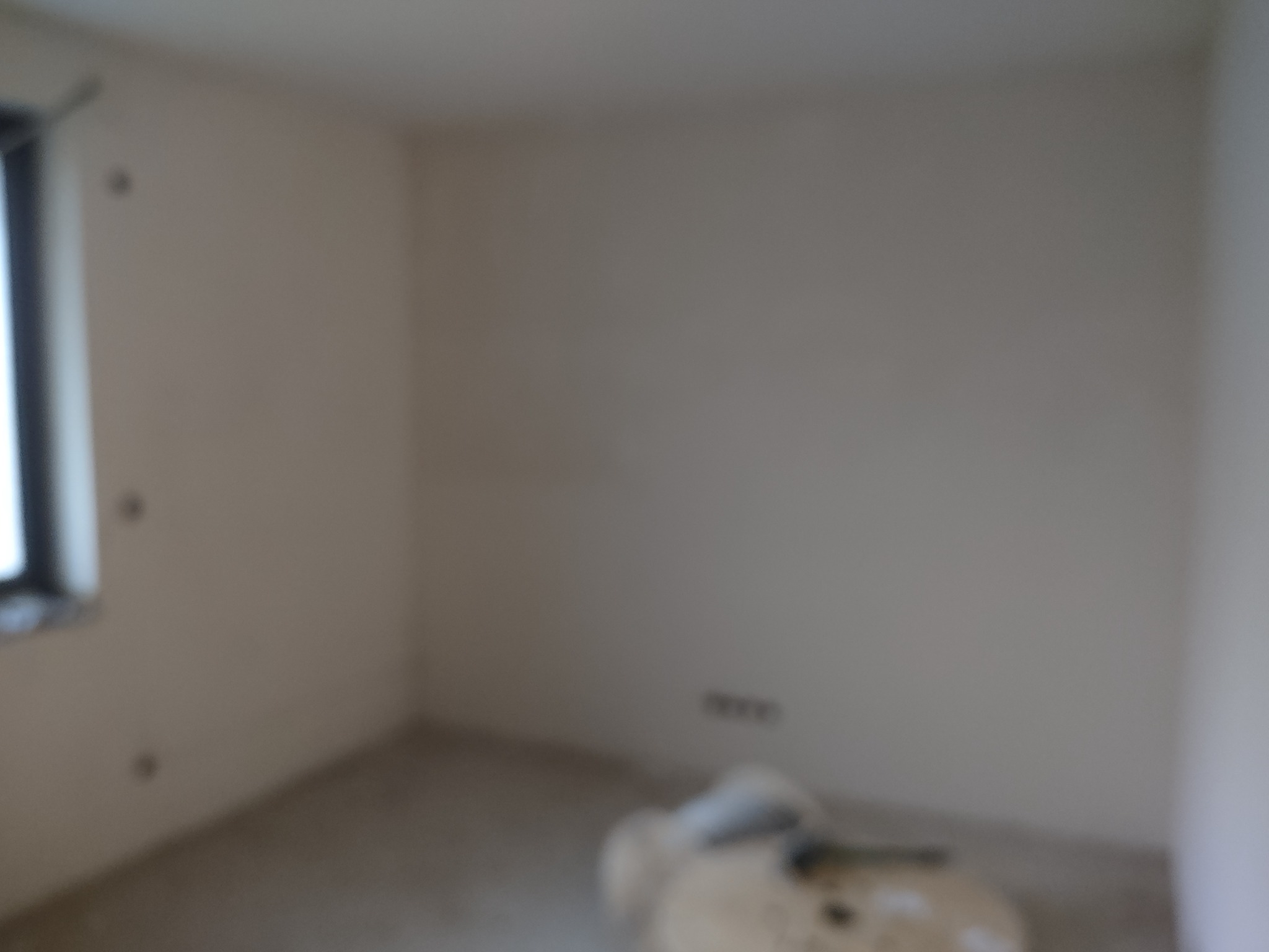 Nierozpoznawalne wnętrze pokoju w trakcie remontu, widoczne ślady po instalacjach elektrycznych na ścianie, częściowo zakryte materiały budowlane na podłodze, słabe oświetlenie.