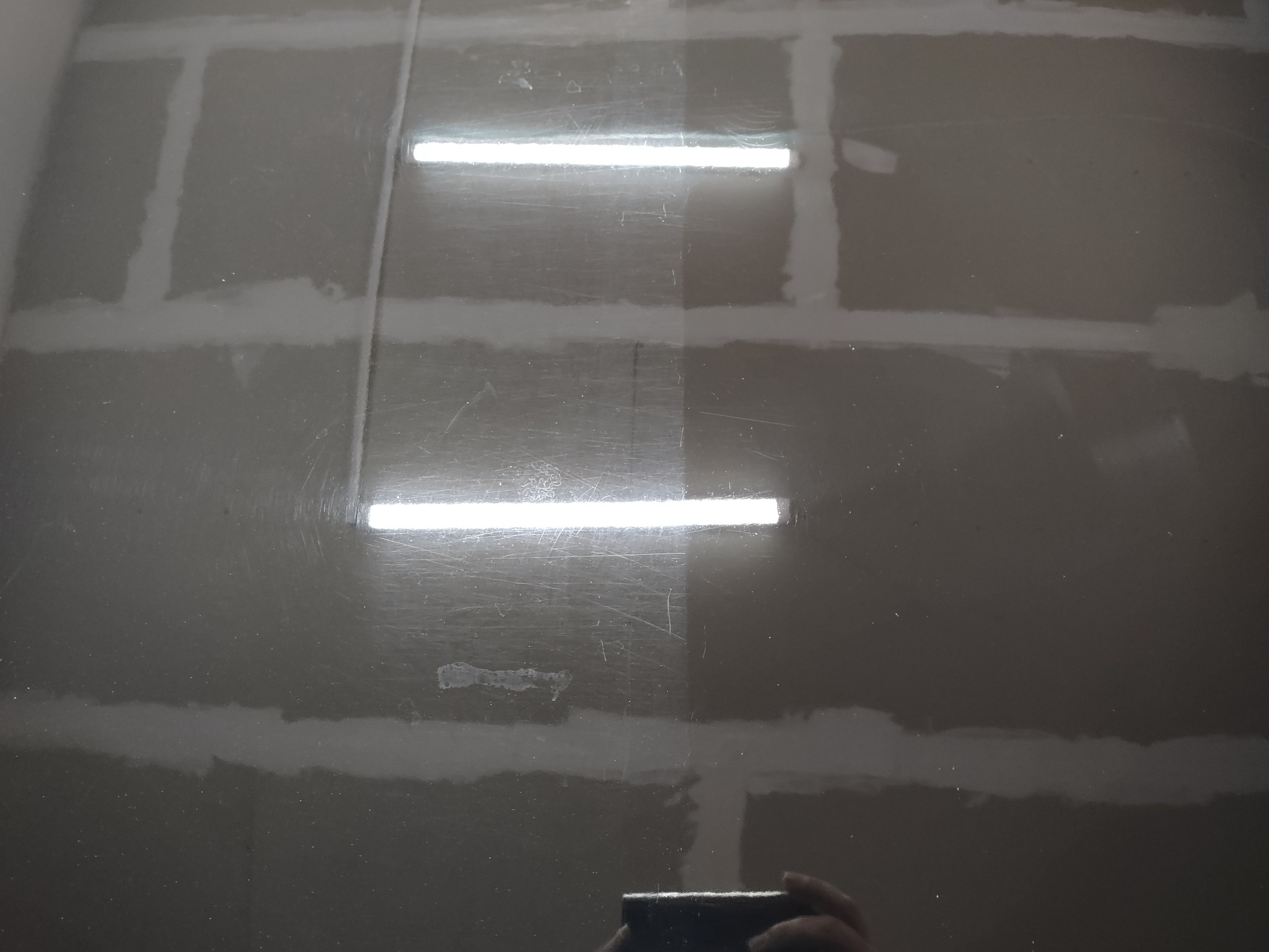 Gładź szpachlowa na ścianie z widocznymi śladami po szlifowaniu, odbijająca światło lamp, z fragmentem odbicia telefonu w dolnej części.