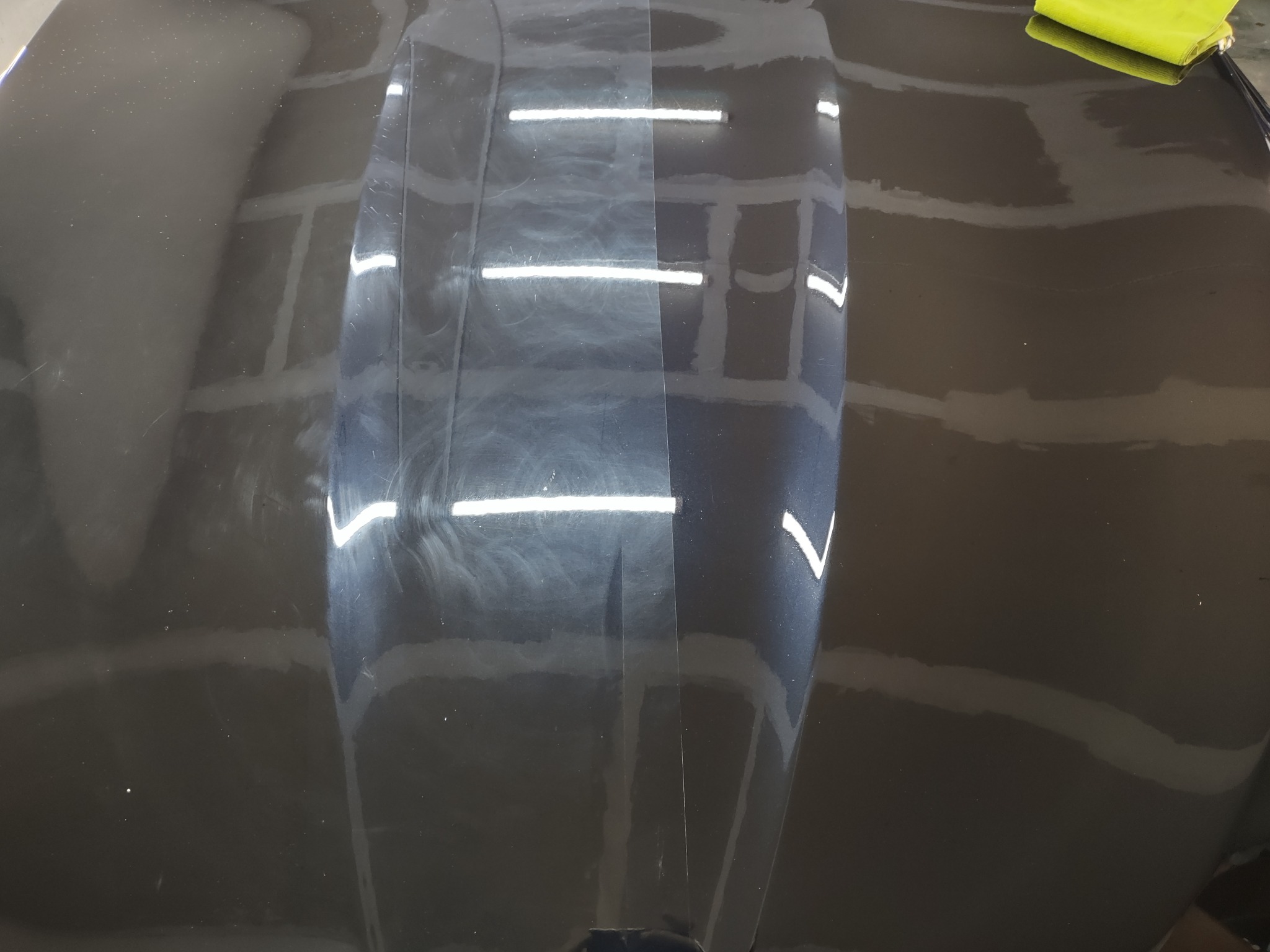 Częściowo wypolerowana, ciemna karoseria samochodu z widocznymi hologramami i zarysowaniami po jednej stronie oraz gładką, odbijającą światło powierzchnią po drugiej. W prawym górnym rogu leży...