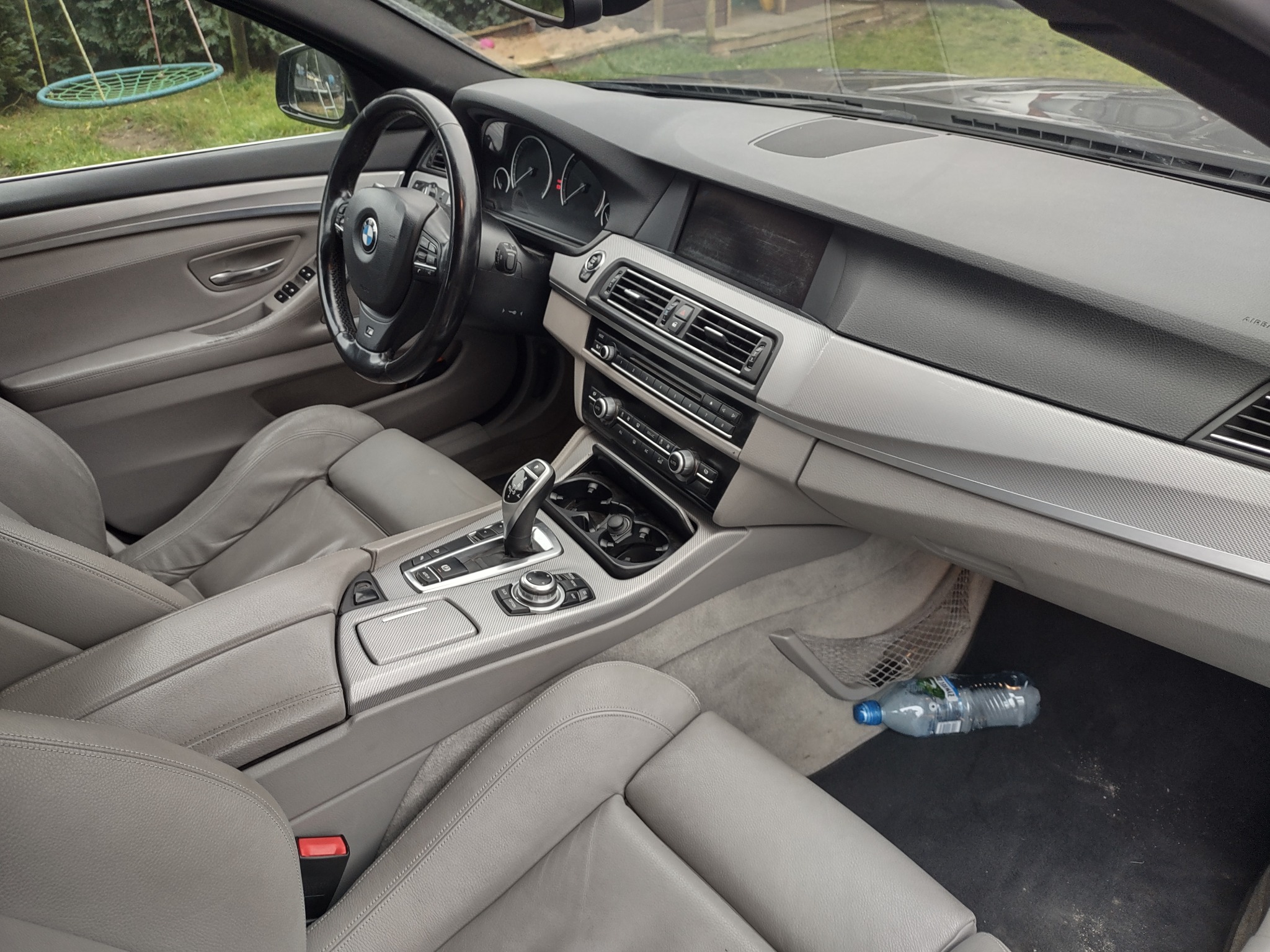 Wnętrze samochodu BMW serii 5 z jasną skórzaną tapicerką, widoczna deska rozdzielcza, kierownica, fotele oraz butelka wody na dywaniku pasażera.