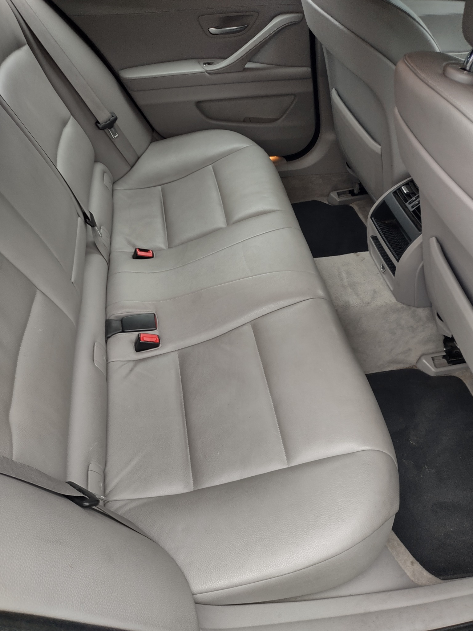 Wnętrze samochodu z widoczną tylną, skórzaną kanapą w jasnym odcieniu szarości, czarnymi dywanikami i kratką wentylacyjną w tunelu środkowym.