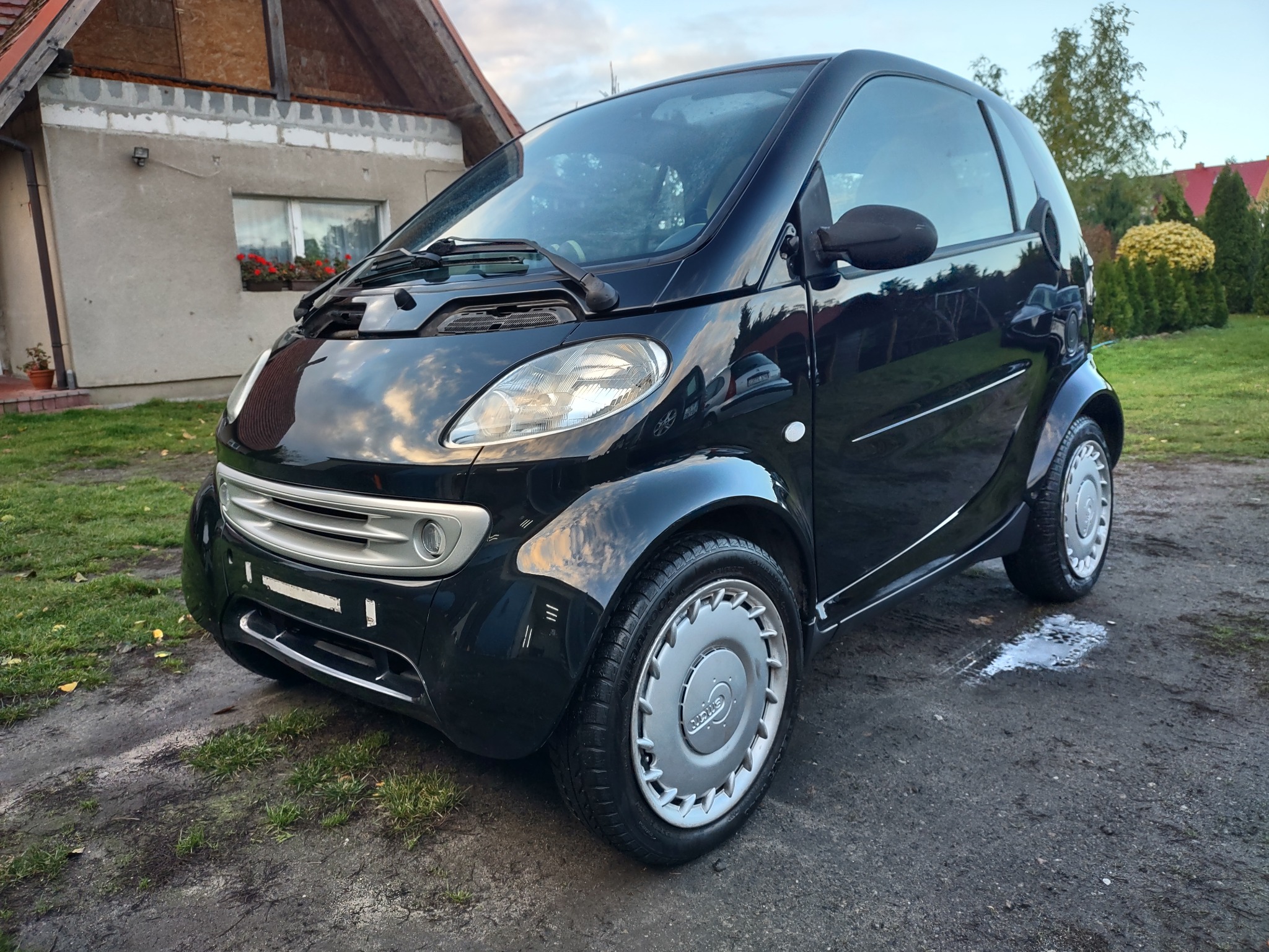 Czarny, miejski samochód Smart zaparkowany na ubitej ziemi przed domem jednorodzinnym z czerwonymi kwiatami w oknie.