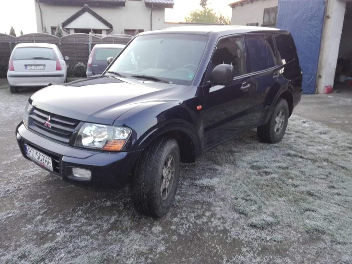 Ciemnoniebieski samochód terenowy Mitsubishi Pajero stoi na zewnątrz na trawie pokrytej szronem, z widocznymi dwoma innymi samochodami i budynkiem mieszkalnym w tle.