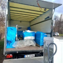 Otwarta paka samochodu dostawczego, załadowana meblami i sprzętem AGD zabezpieczonym folią, widoczna pralka na rampie załadunkowej, tablica rejestracyjna pojazdu.