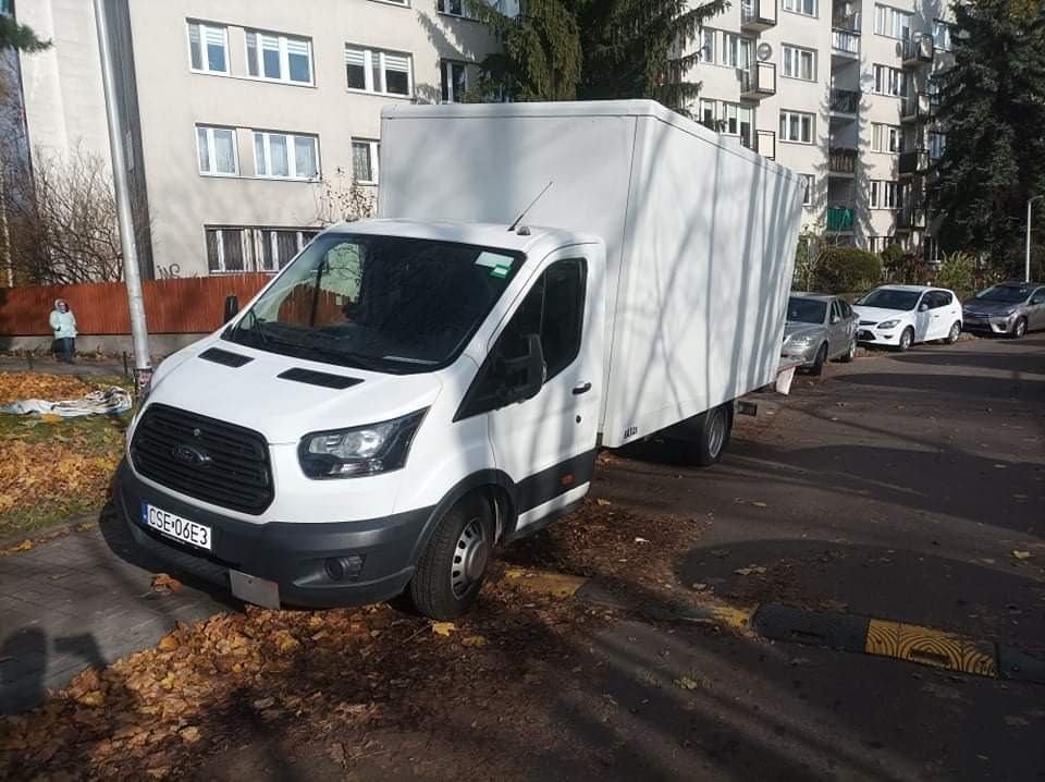 Biały furgon dostawczy marki Ford zaparkowany przy chodniku z opadłymi liśćmi, na tle bloku mieszkalnego i innych zaparkowanych samochodów.