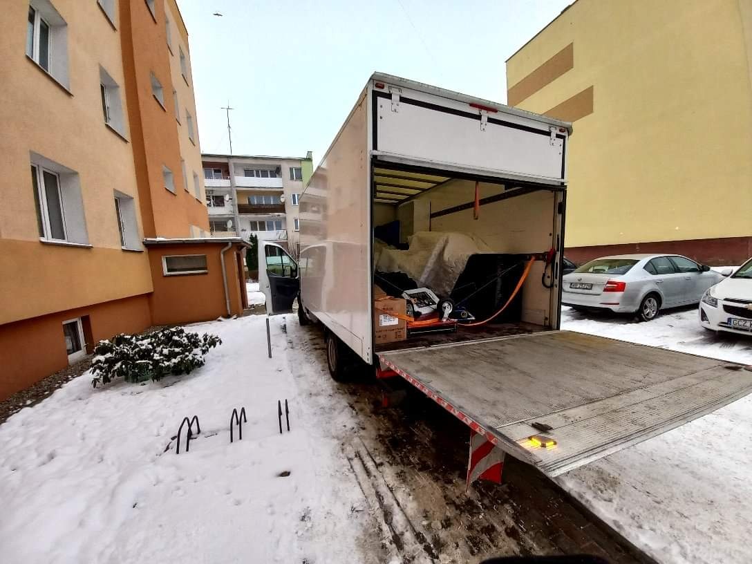 Otwarta skrzynia ładunkowa busa transportowego na śniegu, z widocznym zabezpieczonym ładunkiem wewnątrz, z opuszczoną rampą i zaparkowanymi samochodami osobowymi w tle.