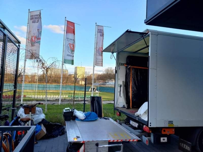 Ciężarówka z otwartą rampą załadowczą, załadowana przedmiotami owiniętymi w czarną folię, na tle miejskiego krajobrazu z rzeką i budynkami, obok stoją rowery i torby.