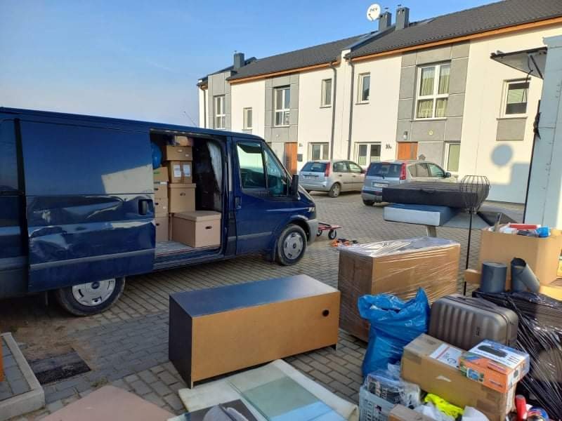 Załadunek mebli i kartonów do niebieskiego busa na tle osiedla mieszkaniowego. Część przedmiotów spakowana i przygotowana do transportu na zewnątrz pojazdu.