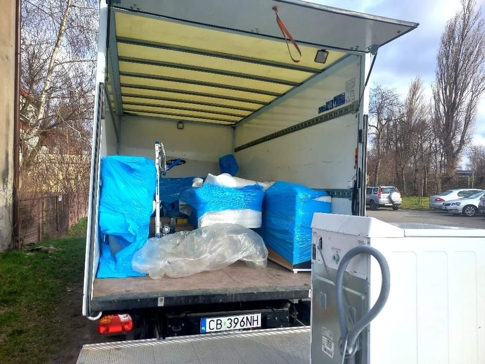 Otwarta paka samochodu dostawczego, załadowana meblami i sprzętem AGD zabezpieczonym folią, widoczna pralka na rampie załadunkowej, tablica rejestracyjna pojazdu.