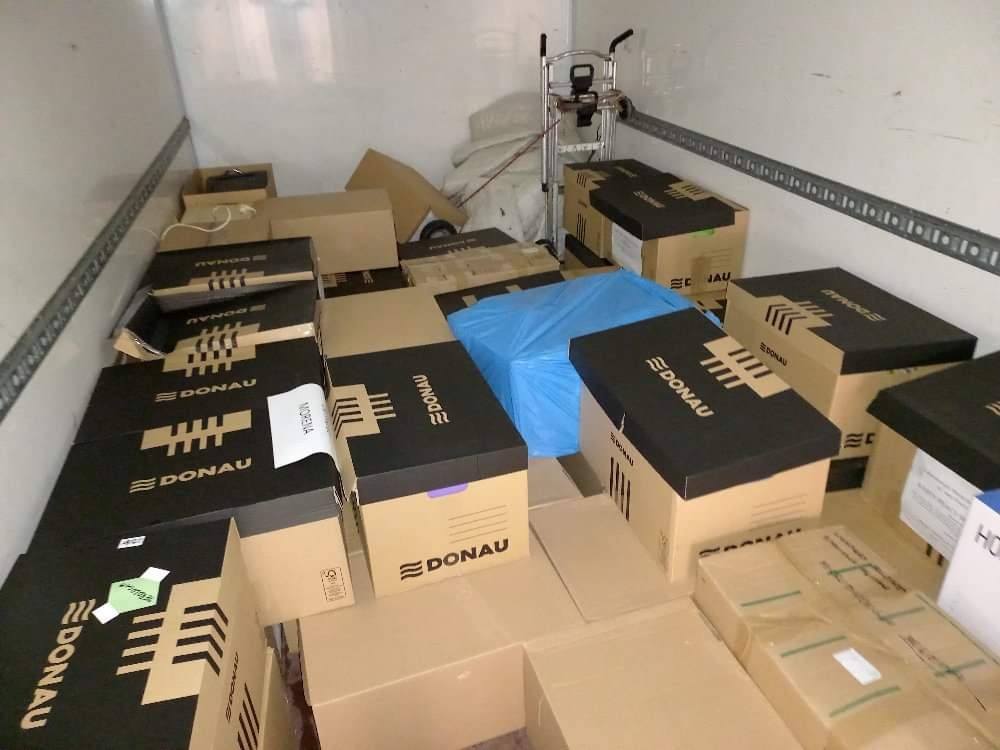 Wnętrze furgonetki wypełnionej kartonami z logo DONAU, częściowo przykrytymi niebieską folią, z wózkiem transportowym w tle.