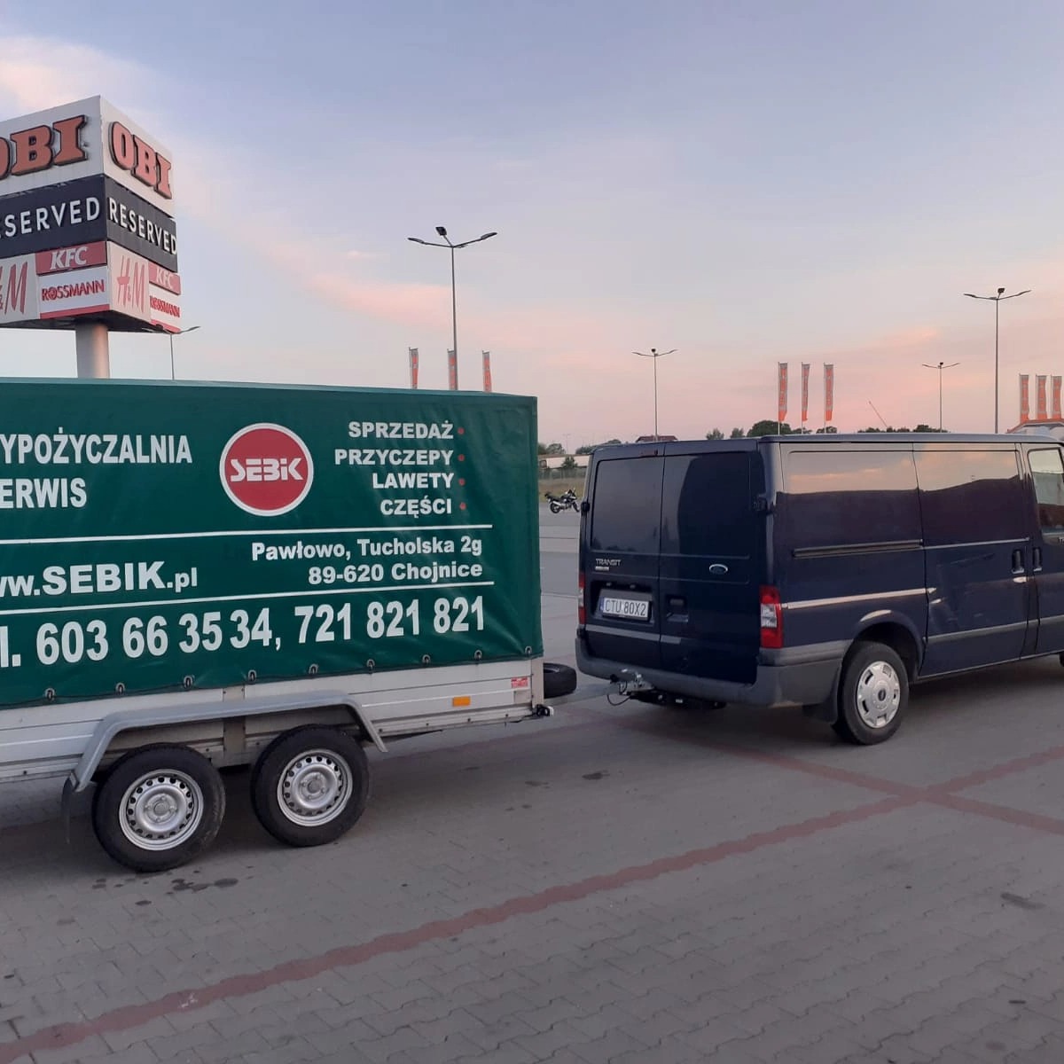 Granatowy bus Ford Transit z przyczepą reklamową firmy Sebik na parkingu przed centrum handlowym z szyldami OBI, KFC i Rossmann.