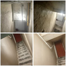 Szym-bud - Klatka schodowa w trakcie prac wykończeniowych; ściany i schody pokryte świeżym tynkiem, widoczne elementy instalacji i drzwi.