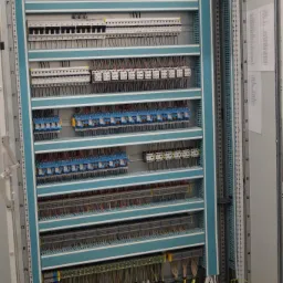 Otwarta szafa sterownicza z licznymi zabezpieczeniami, wyłącznikami i okablowaniem, widoczne oznaczenia na przewodach.