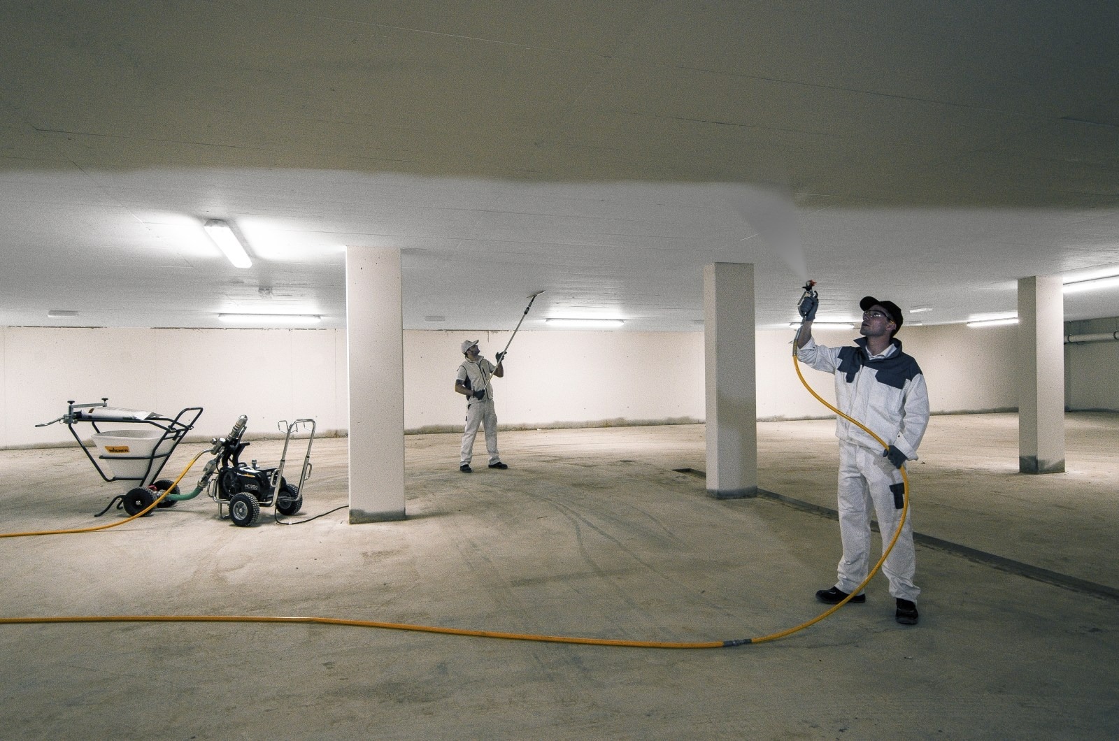 Dwóch pracowników w kombinezonach roboczych, używających pistoletów natryskowych do pokrywania sufitu w garażu podziemnym, z widocznym sprzętem do natrysku farby i żółtym wężem na posadzce.
