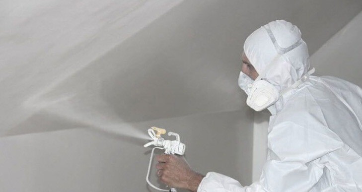 Pracownik w kombinezonie ochronnym natryskuje ścianę i sufit agregatem malarskim, widoczna mgiełka farby.