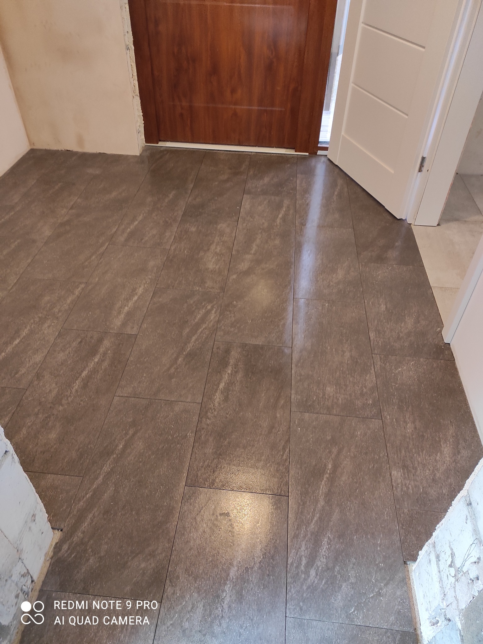 Ułożona, brązowo-szara podłoga z płytek imitujących kamień, widok z góry, z fragmentem białej i brązowej futryny drzwi. W tle surowy fragment ściany.