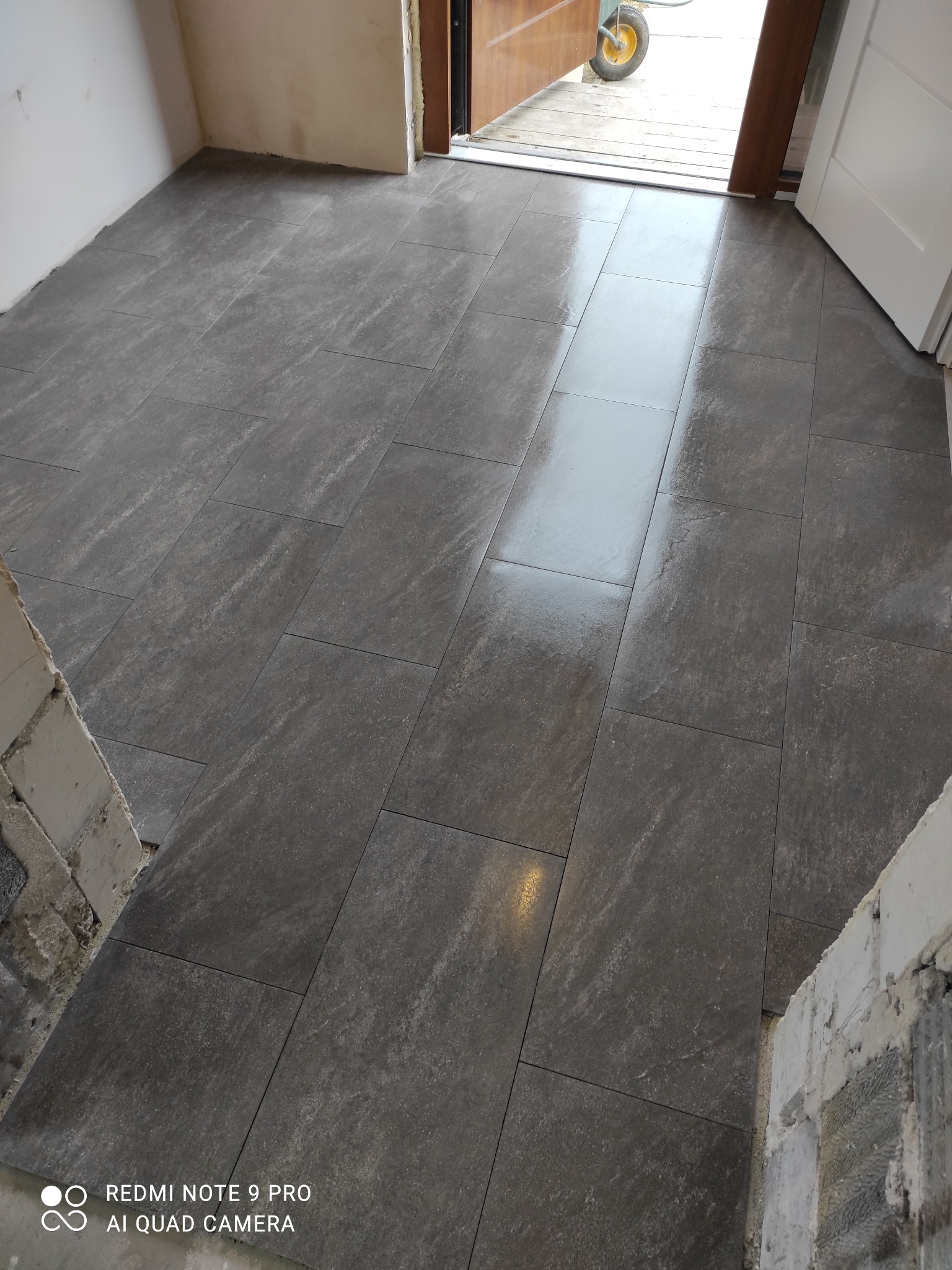 Ułożona, szara, prostokątna terakota podłogowa w korytarzu. Widoczne niedokończone fragmenty ścian. W tle otwarte drzwi z widokiem na zewnątrz.