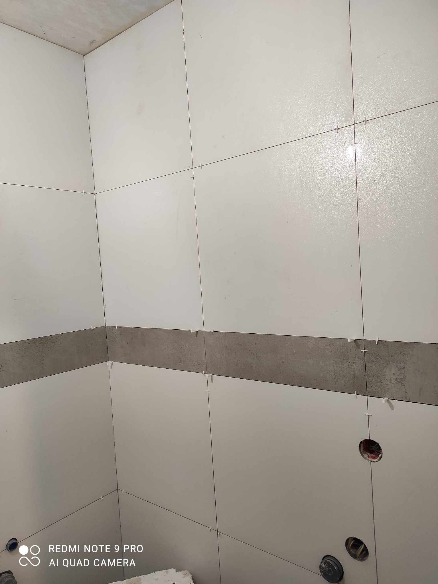 Ściana w trakcie układania białych płytek z szarą listwą dekoracyjną, widoczne krzyżyki dystansowe i otwory na instalacje. Surowy betonowy sufit.