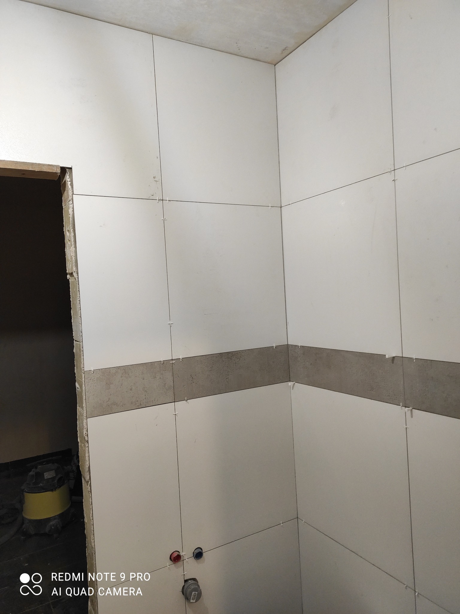 Układanie białych i szarych płytek w łazience w Choinach. Widoczne krzyżyki dystansowe i fragment odkurzacza. Świeżo położona glazura na ścianach.