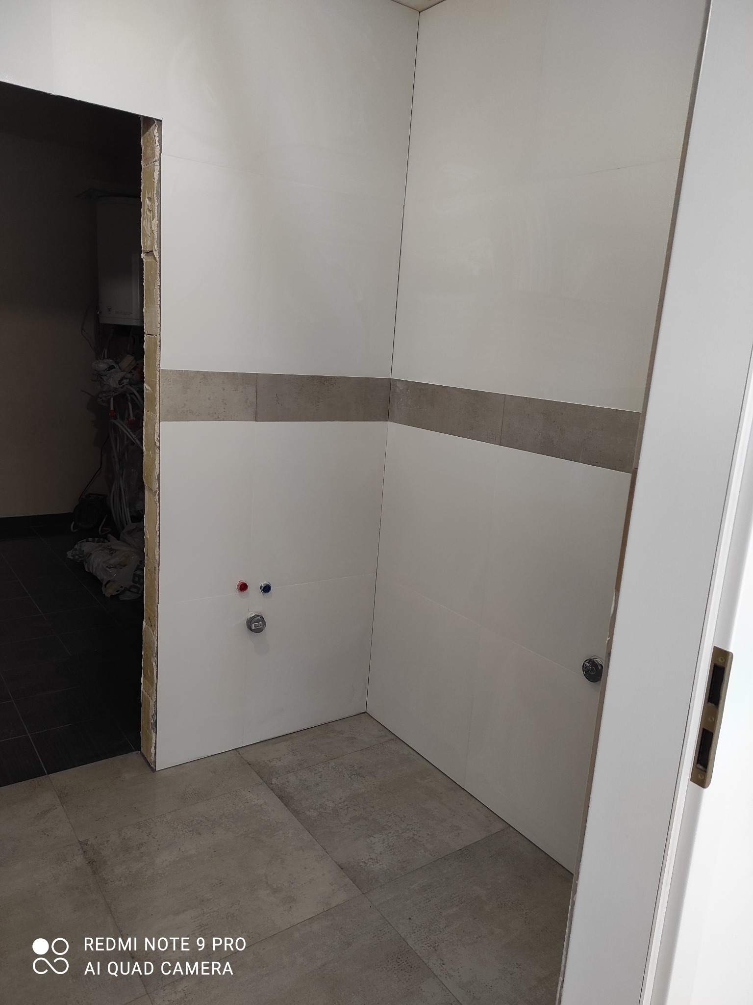 Łazienka w trakcie remontu: białe płytki ścienne z szarym pasem, szara podłoga, widoczne instalacje hydrauliczne i otwarte przejście do innego pomieszczenia.