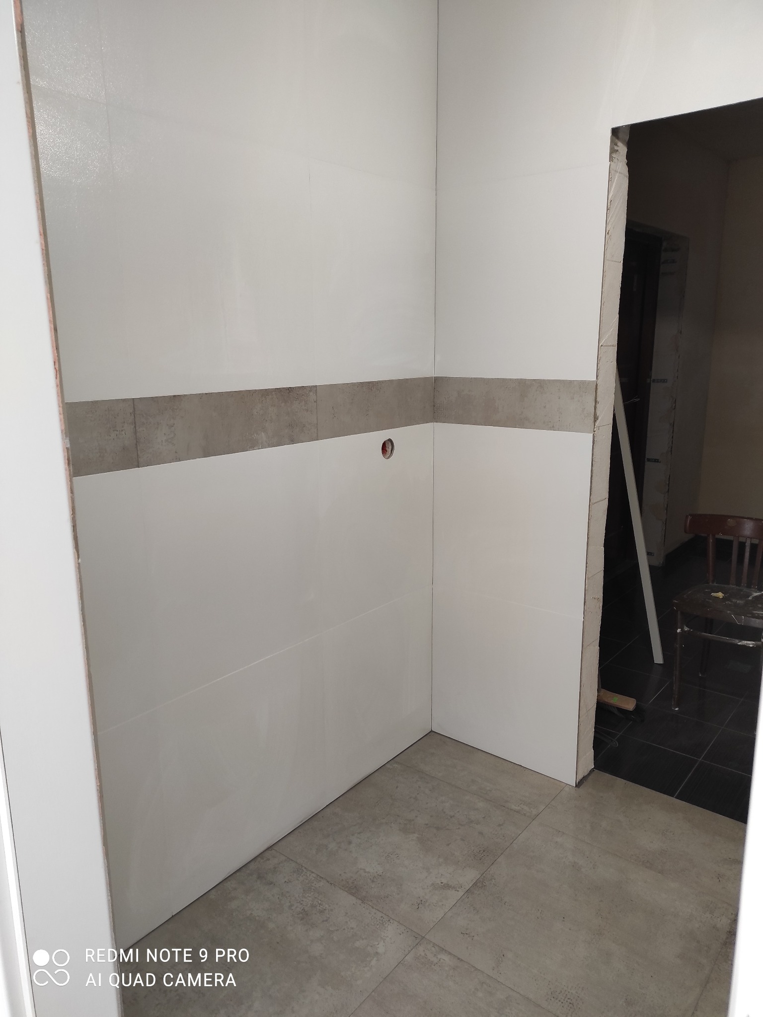 Wykończona ściana w łazience z białymi płytkami i szarym pasem dekoracyjnym. Na podłodze szare płytki. Widoczny fragment drzwi i krzesło w tle.