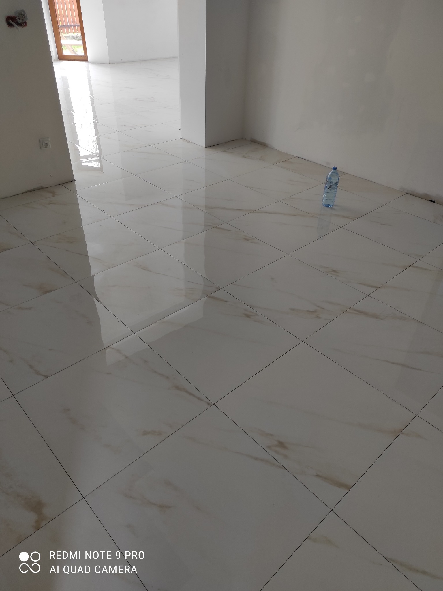 Położona, błyszcząca, beżowo-biała podłoga z płytek w nowym, jasnym pomieszczeniu. W tle widoczne okno i butelka wody. Minimalistyczny efekt wykończenia.