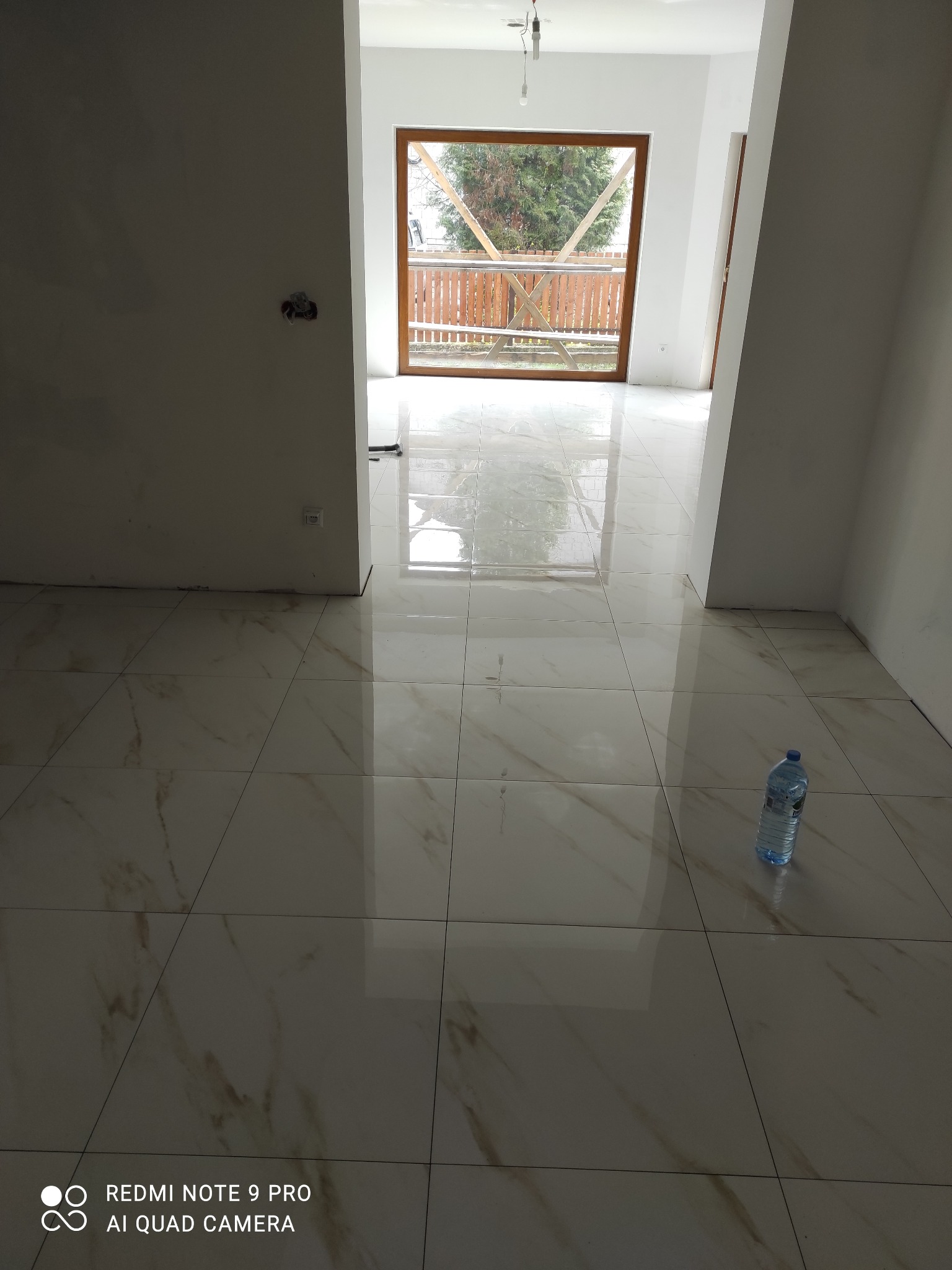 Położone płytki podłogowe w jasnym odcieniu z marmurkowym wzorem w nowym wnętrzu. Widoczna butelka wody. Okno balkonowe z widokiem na zewnątrz.