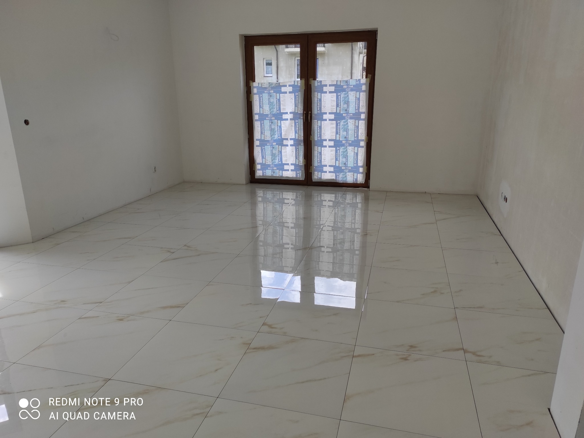 Błyszczące, kremowe płytki podłogowe z delikatnym wzorem żyłkowania w pustym pokoju. Okno zabezpieczone folią ochronną. Świeżo położona podłoga.