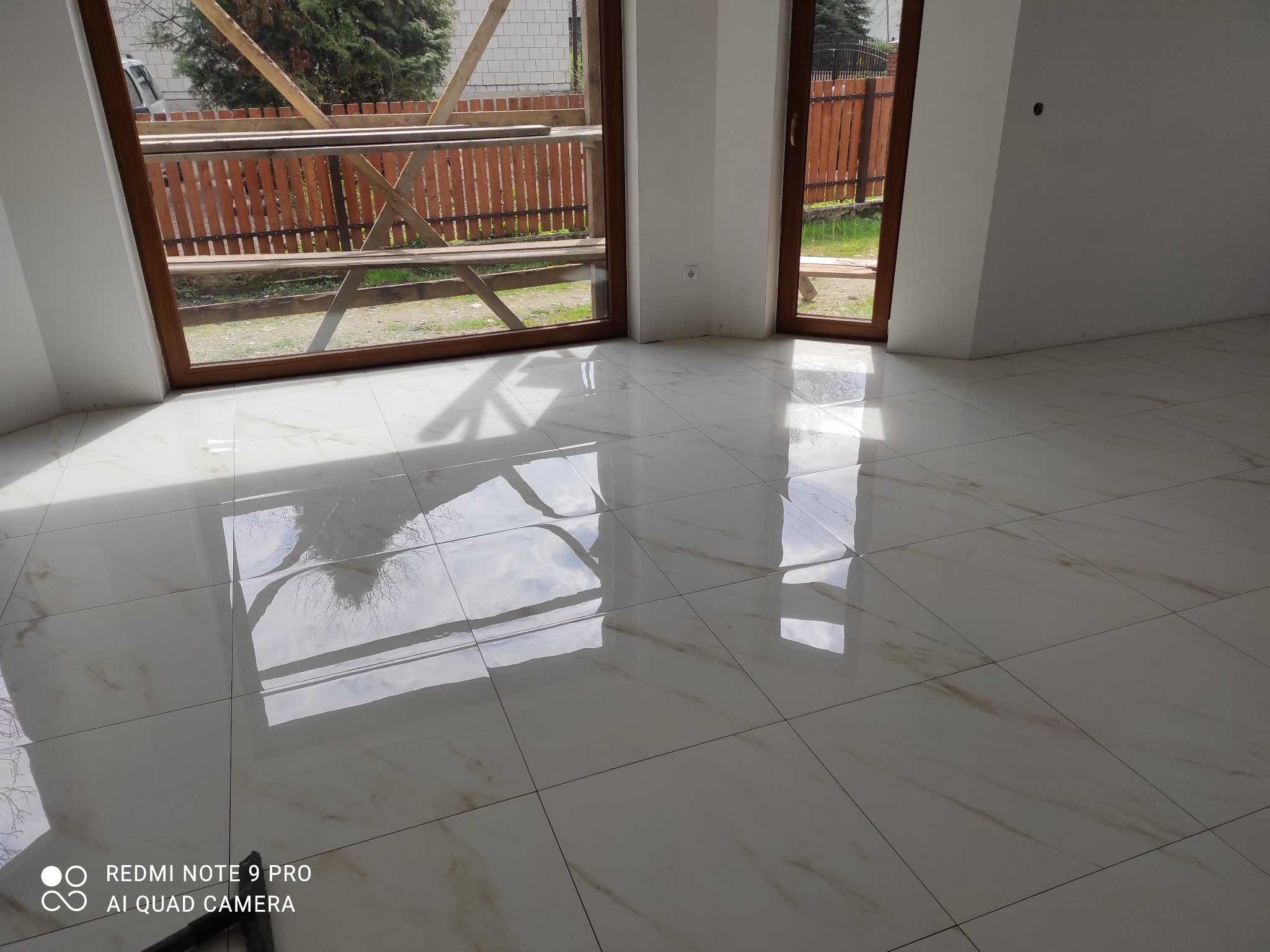 Błyszcząca, jasna podłoga z płytek w nowym wnętrzu. Widoczne duże okna z drewnianymi ramami i odbicia w podłodze. Minimalistyczny styl, świeżo po remoncie.