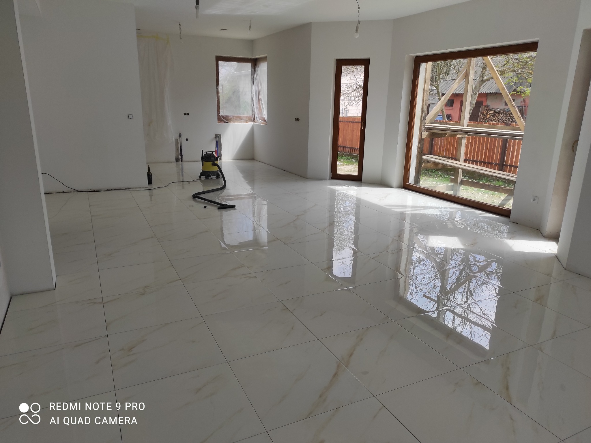 Błyszcząca, kremowa podłoga z płytek imitujących marmur w przestronnym, jasnym pomieszczeniu po remoncie. Widoczne okna i drzwi balkonowe, odkurzacz przemysłowy.