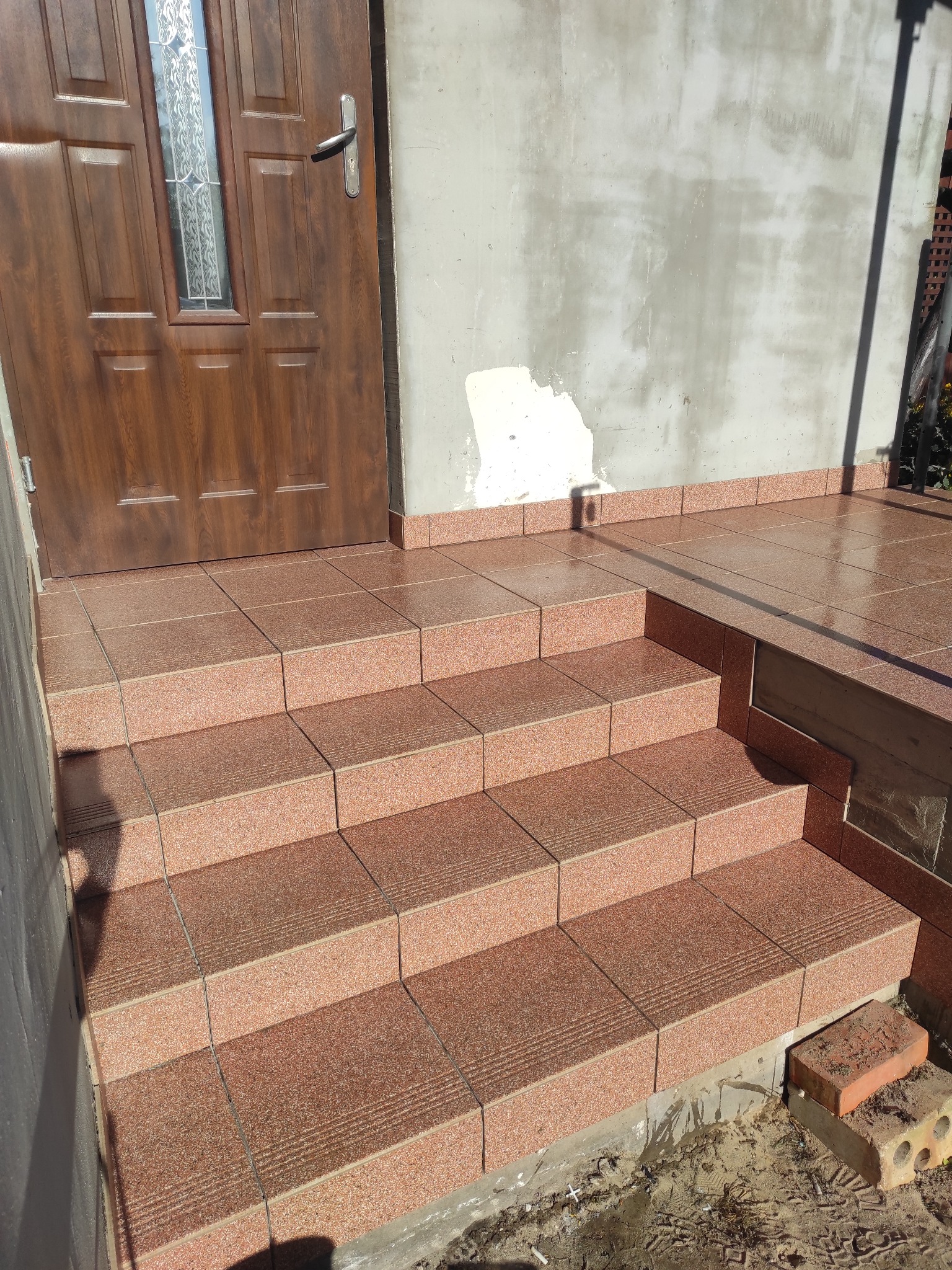 Wykończone płytkami schody wejściowe do domu z brązowymi drzwiami. Widoczne detale: fuga, faktura płytek, fragment elewacji w trakcie prac wykończeniowych.