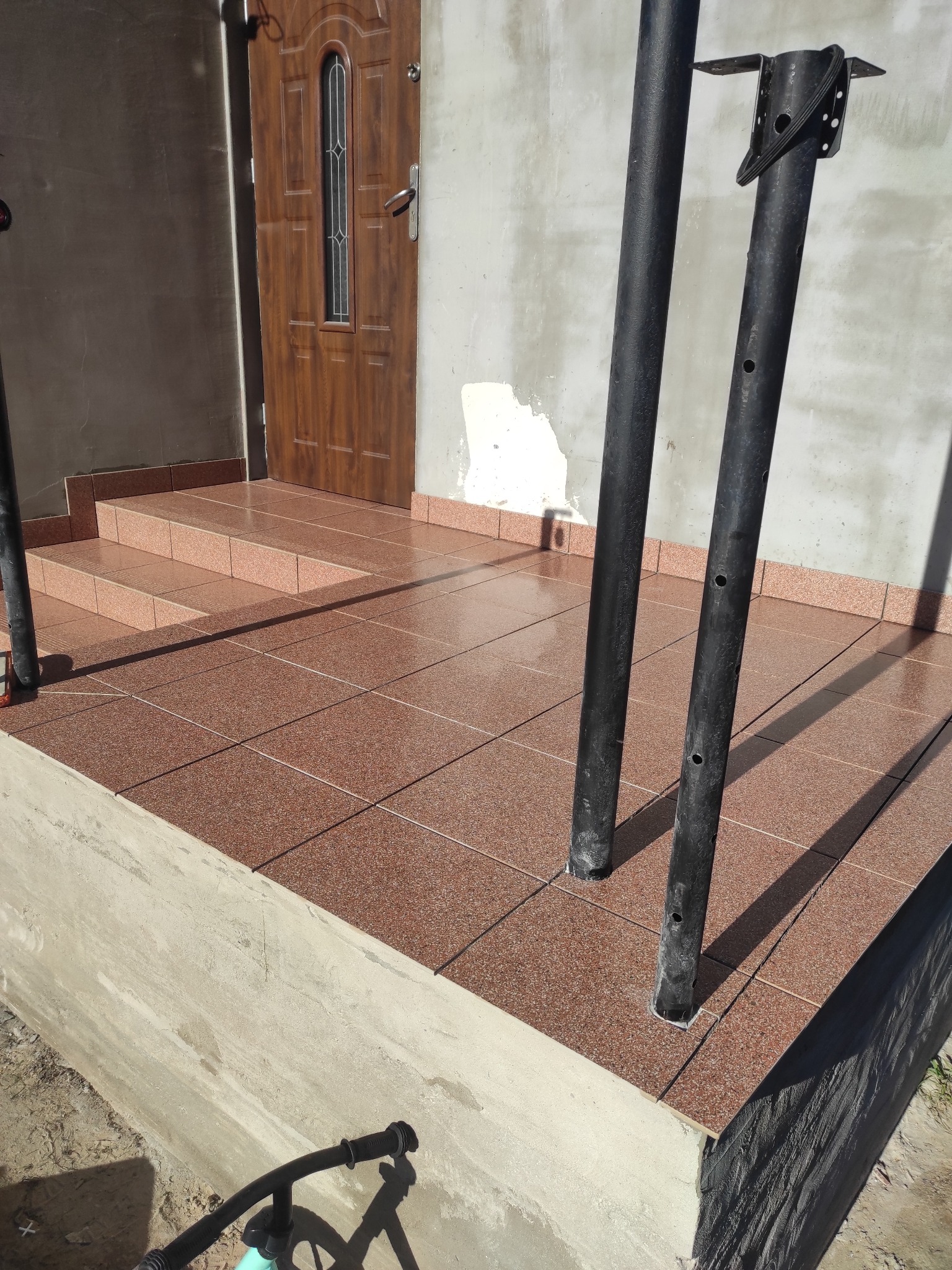 Wykończony taras z brązowych płytek, widoczne schody wejściowe i fragment drzwi wejściowych. Na pierwszym planie czarne podpory. Słoneczny dzień.