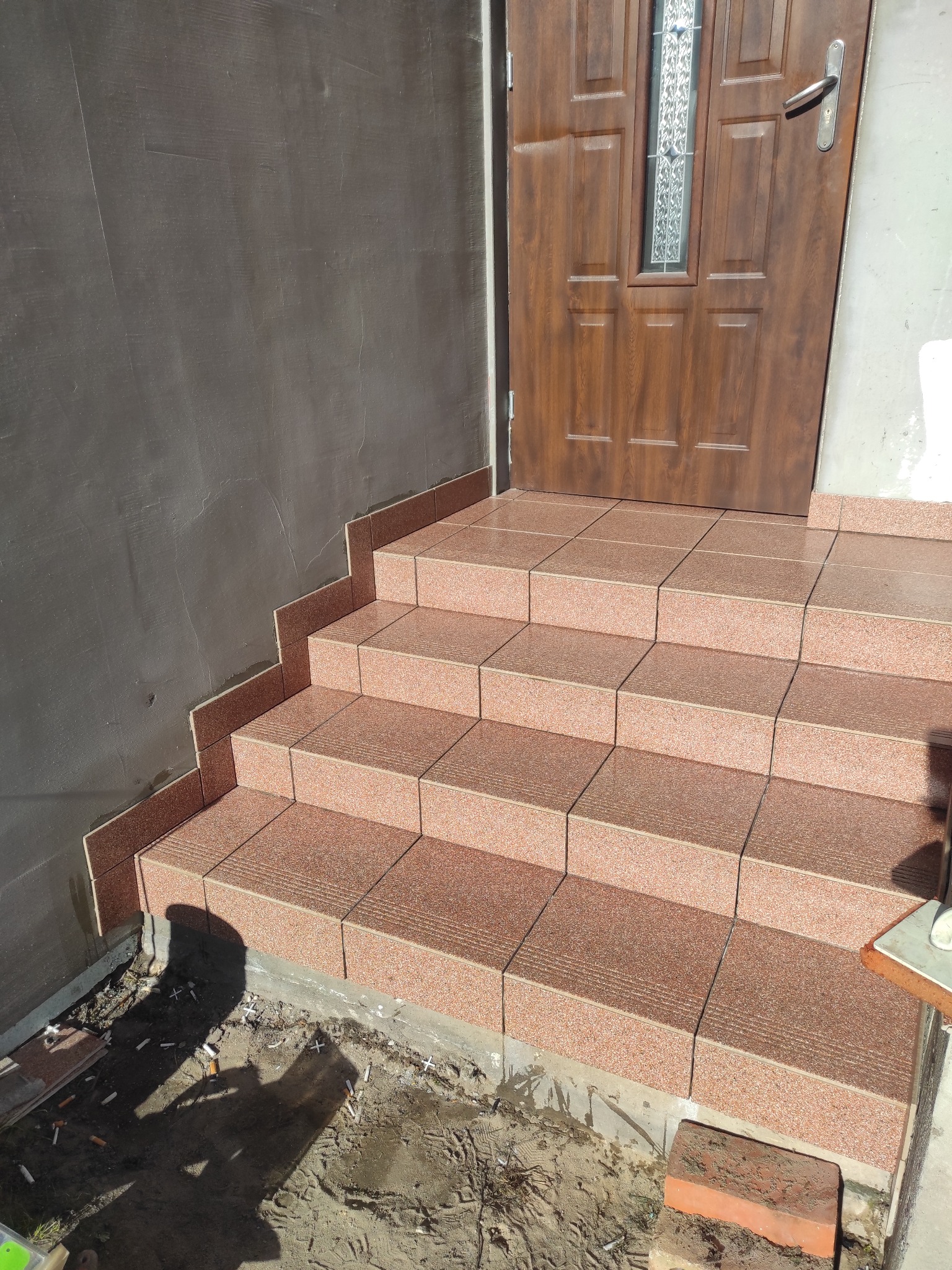 Wykończone płytkami schody wejściowe w Choinach, prowadzące do brązowych drzwi. Widoczne detale fug i struktura płytek. Ściana obok schodów częściowo otynkowana.