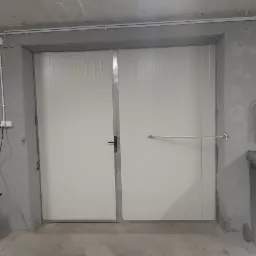 Dwuskrzydłowa, biała, wewnętrzna brama z metalowymi wzmocnieniami i poprzeczką zabezpieczającą, zamontowana w surowym, betonowym pomieszczeniu.