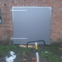 Szara dwuskrzydłowa brama z metalowymi zawiasami zamontowana w czerwonej ceglanej ścianie, widoczny wózek paletowy i resztki folii na ziemi.