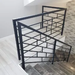 Nowoczesna, czarna balustrada schodowa z geometrycznym wzorem, kontrastująca z szarymi schodami i ścianą z dekoracyjnej cegły.