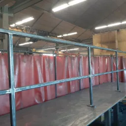 Surowa, metalowa konstrukcja balustrady w warsztacie, z czerwonymi zasłonami spawalniczymi w tle, widoczne spawy i szlifowane łączenia.