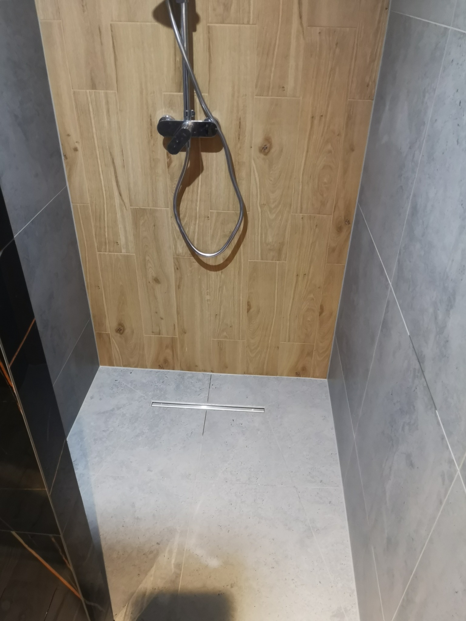 Kabina prysznicowa wykończona płytkami imitującymi drewno i szarym gresem, z odpływem liniowym i chromowaną armaturą.