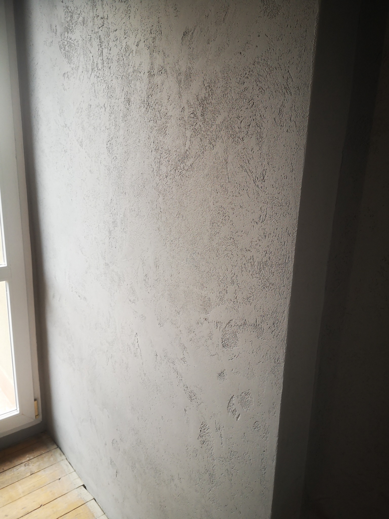 Ściana wewnętrzna pokryta szarą, teksturowaną farbą dekoracyjną, widoczny fragment okna z białymi ramami i drewniana podłoga z desek w odcieniach beżu, z wyraźną fakturą na ścianie.