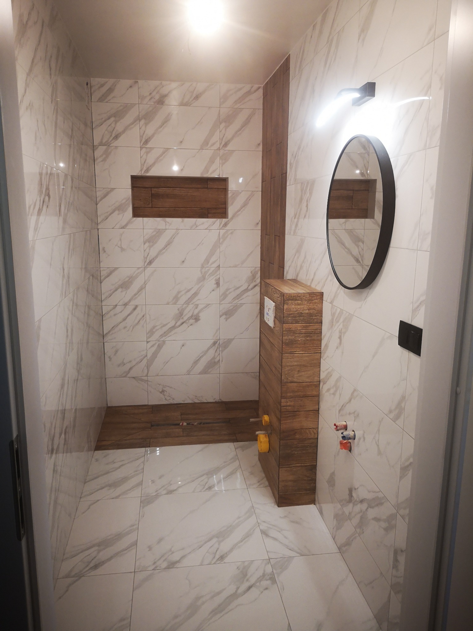 Nowoczesna łazienka z marmurowymi płytkami, drewnianymi akcentami i okrągłym lustrem. Podtynkowa toaleta z drewnianą obudową, widoczne przyłącza wodne z kolorowymi zaworami.