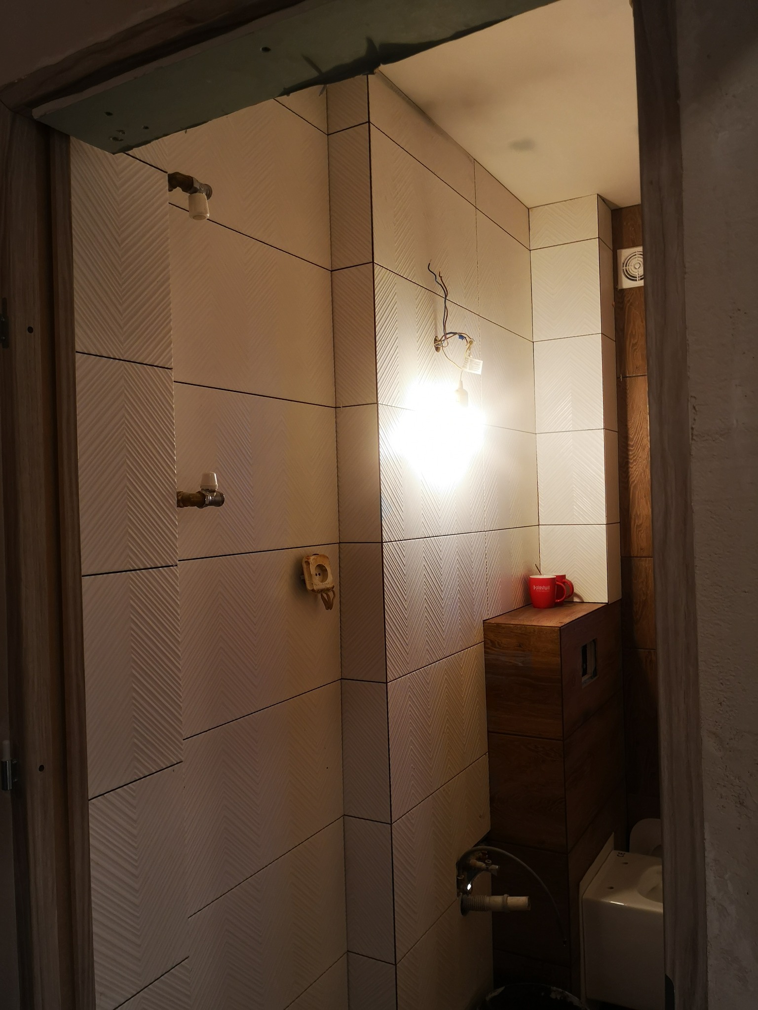 Łazienka w trakcie wykańczania: kremowe płytki z delikatnym wzorem jodełki na ścianach, widoczne przyłącza wodne, drewniana szafka z czerwonym kubkiem na blacie, surowy stan wykończenia sufitu...