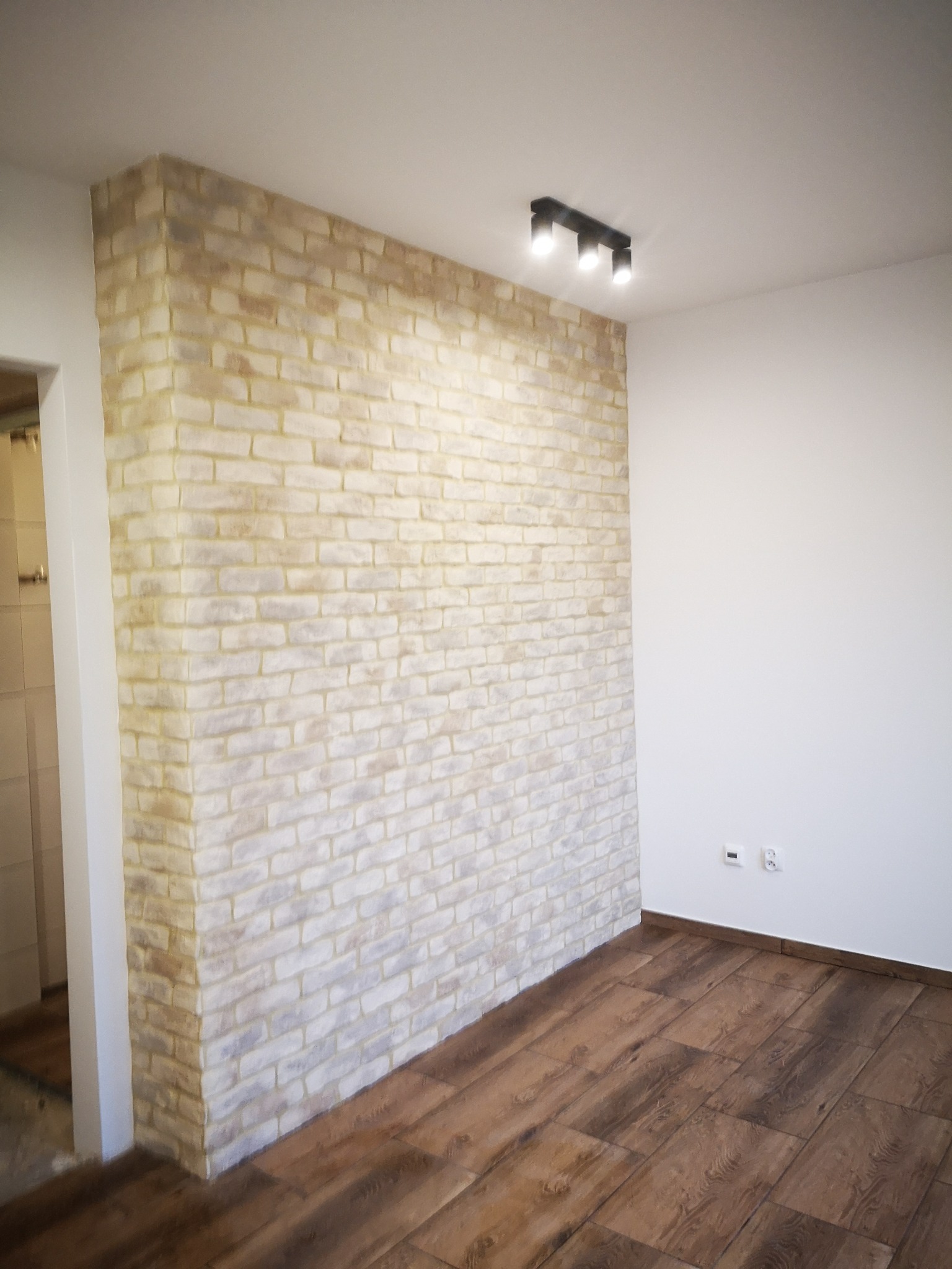 Ściana z dekoracyjnej cegły w odcieniach bieli i beżu, oświetlona przez czarną lampę sufitową z trzema reflektorami, z podłogą z paneli imitujących drewno w nowocześnie wykończonym wnętrzu.