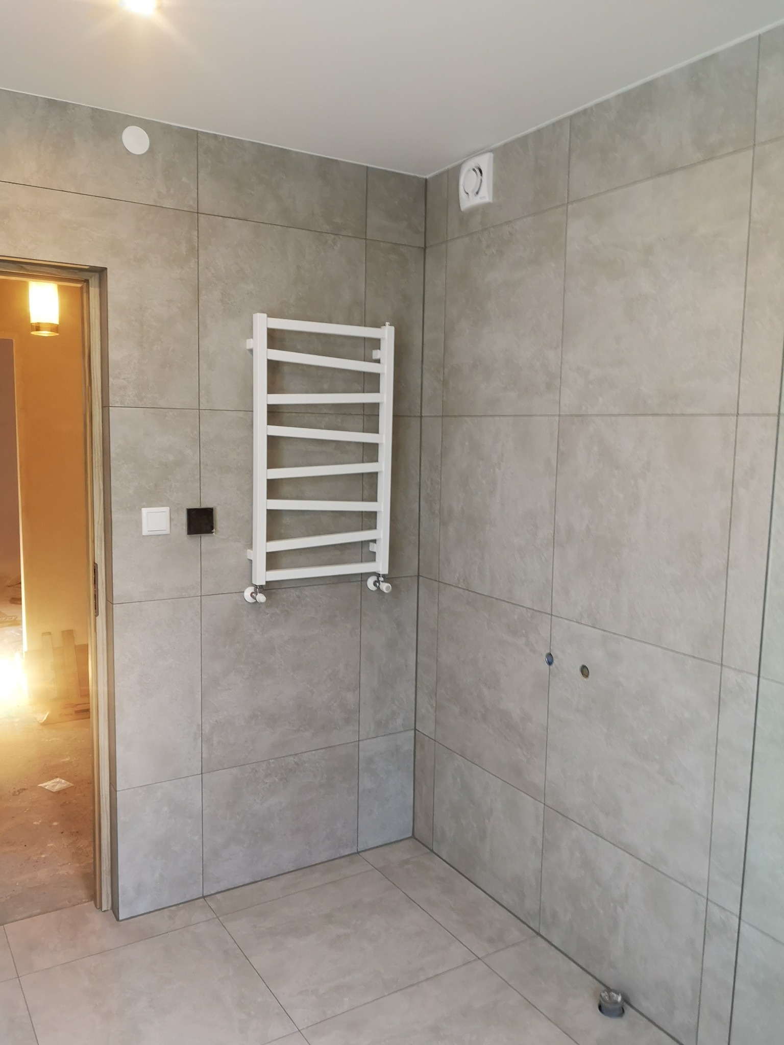 Jasnoszara, kwadratowa płytka ceramiczna na ścianie w łazience, z zamontowanym białym grzejnikiem drabinkowym i widocznymi otworami na baterię, podłoga również wyłożona płytkami, widoczny fragment...