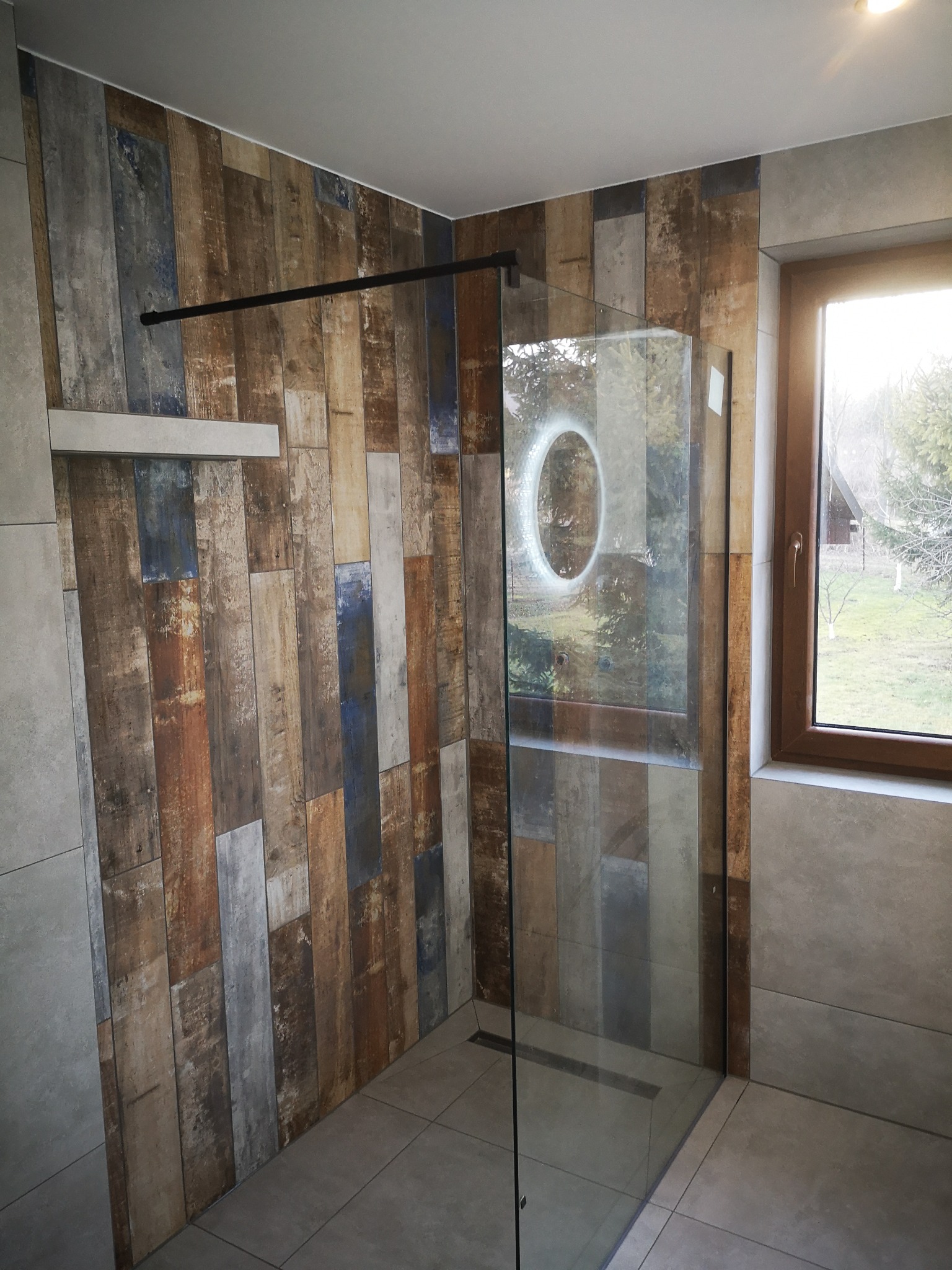 Nowoczesna kabina prysznicowa z szybą z okrągłym wycięciem, obłożona płytkami imitującymi drewno w odcieniach brązu, beżu i niebieskiego, z widokiem na ogród przez okno.