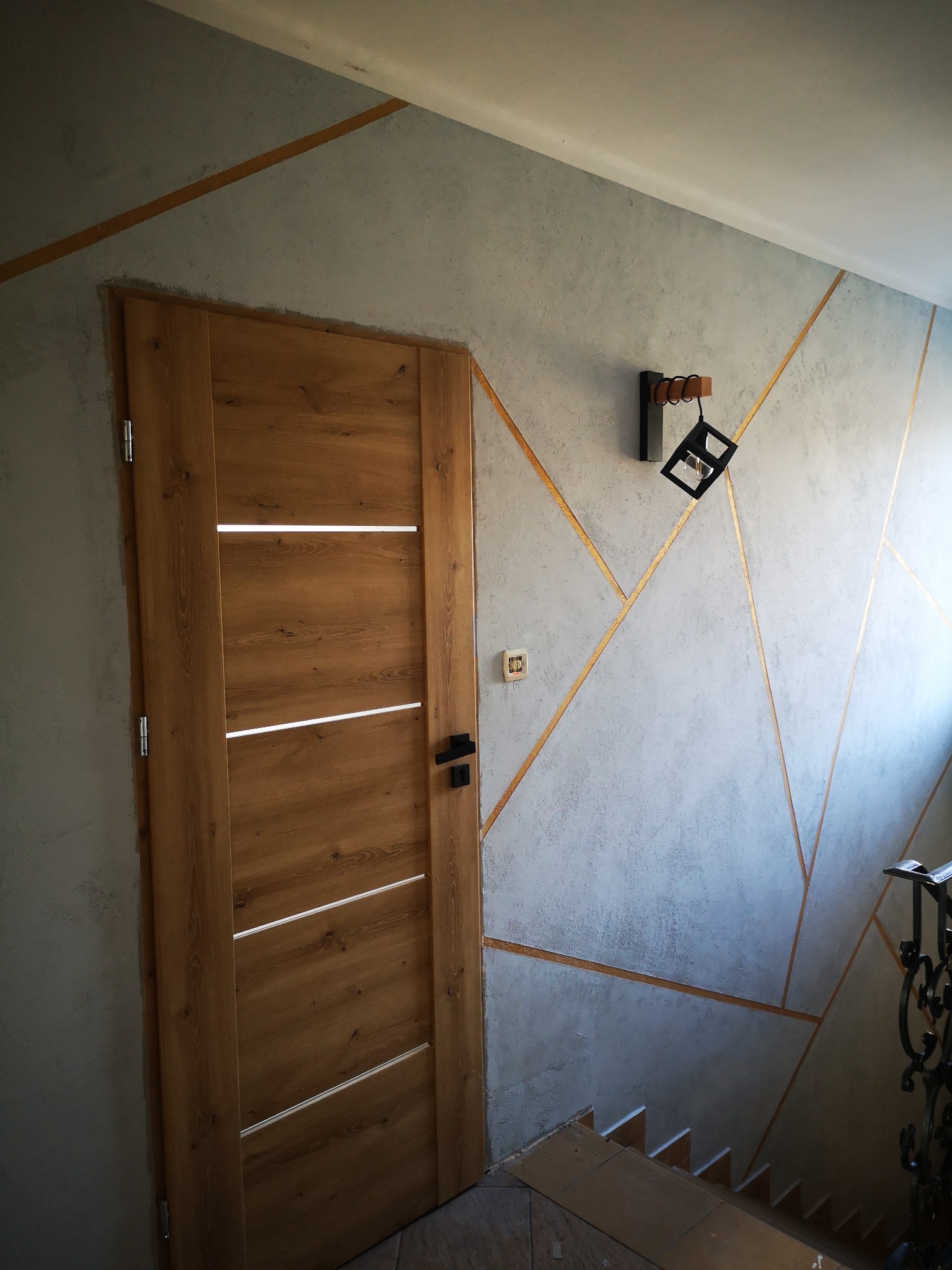 Nowoczesne wnętrze klatki schodowej z drewnianą okleiną drzwi, dekoracyjną szarą ścianą z geometrycznymi złotymi akcentami i designerską lampą sufitową.