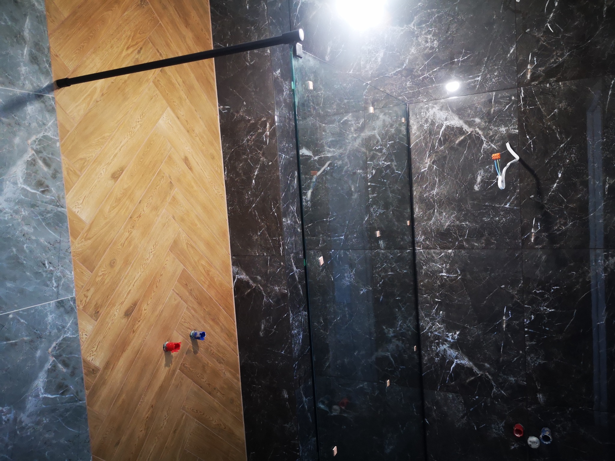 Wnętrze łazienki w trakcie wykańczania: ściana z płytek imitujących ciemny marmur z białymi żyłami, fragment ściany wyłożonej drewnopodobnymi panelami w jodełkę, widoczne przyłącza wodne...