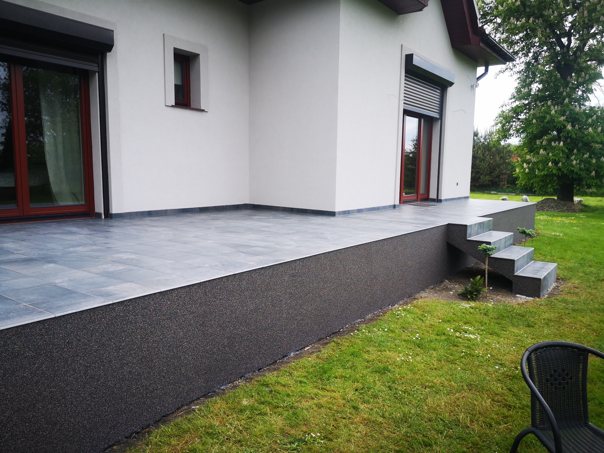 Nowoczesny taras domu jednorodzinnego z szarymi płytkami i schodami, wykończony ciemnym, teksturowanym materiałem na cokole, widok na zadbany trawnik i kwitnące drzewo.